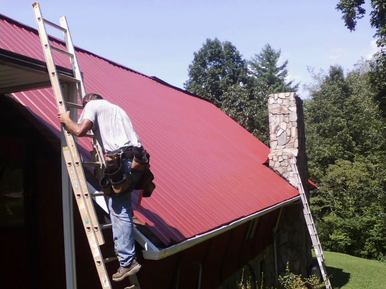 Roofer on a ladder beside a red metal roof and stone chimney — Soffit & Fascia Repair by Roofing Brothers in Iron Station